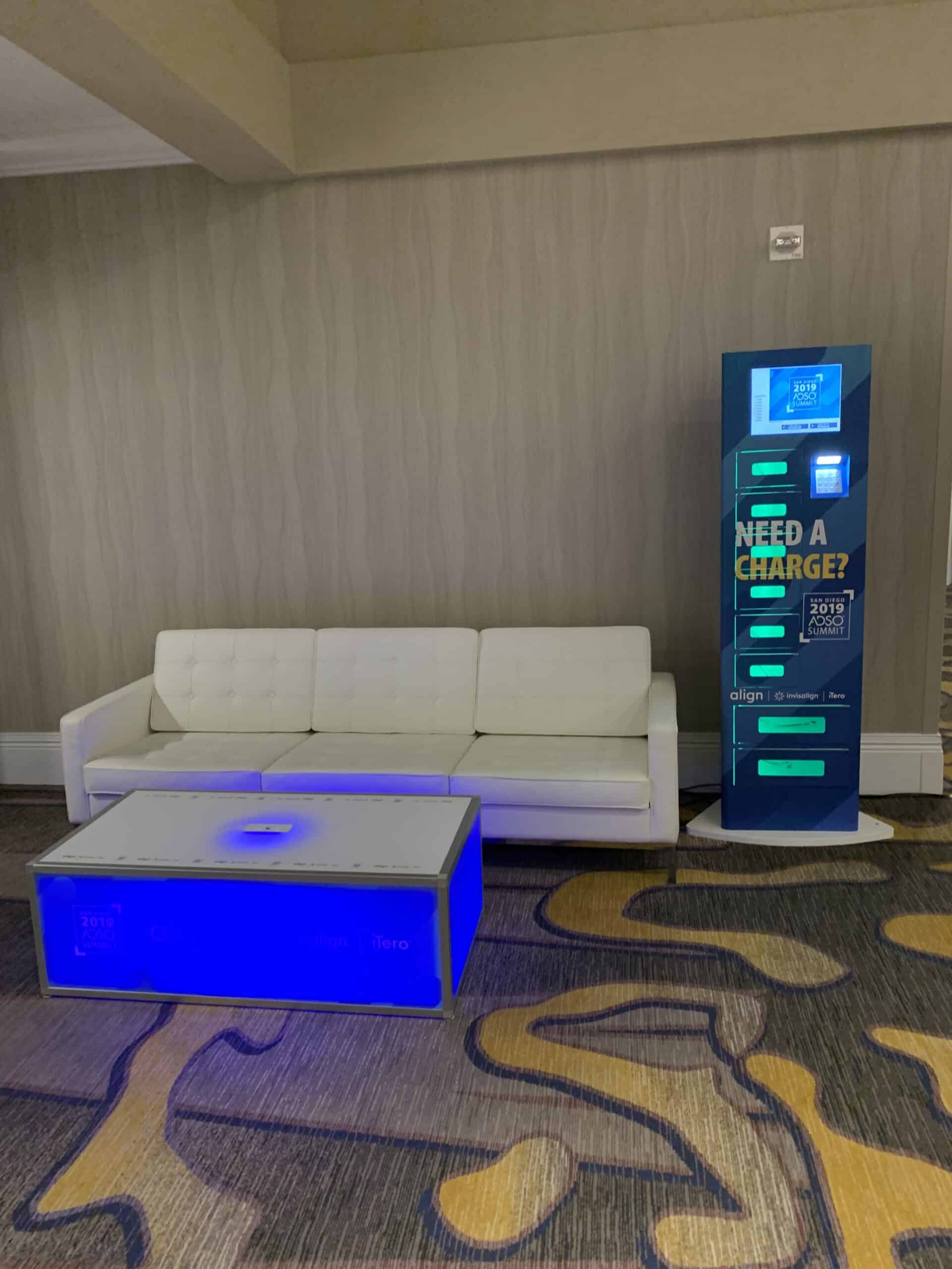 Branded charging locker tower beside a white sofa and illuminated charging coffee table at a conference